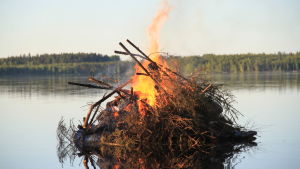 Midsommarbrasa. En midsommarbrasa på en sten ute i vattnet. I bakgrunden en strandkant med träd.