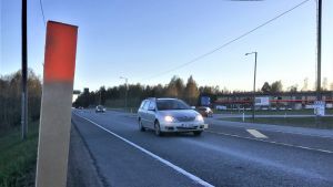 En av käpparna som nu syns vid vägrenarna och på åkrarna invid korsningen i Läpp. Korsningen ses här österifrån. Bilen i förgrunden kommer från Ekenäs- och Hangöhållet. Avtaget till Karis och Pojo (västra omfartsvägen eller väg 111) syns till höger. En T-korsning där personbilar kör åt olika håll. Affärsbyggnader syns i bakgrunden. Asfalterad väg, skymning.
