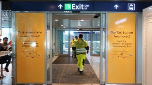 Byggnadsarbeten utanför terminal två har lett till att taxibilarna nu väntar vid ingången på första våningen. Byggarbetare går med ryggen vänd mot kameran ut från Helsingfors-Vanda flygplats. På sidan av skjutdörrarna står texten "Taxistationen har flyttat!"