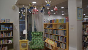 En av många läshörnor i Hangö bibliotek. Vill man vila ögonen och lyfta blicken från boken kan man ta en titt upp i taket och beundra tygdockor som ser ut som sagoälvor. En länstol och ett litet bord med bordslampa i ett bibliotek. Läshörna. I taket hänger tygdockor som ser ut som älvor.