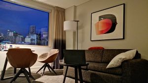Så här ser hotellrummet ut där Pontus Johansson har tillbringat snart 14 dagar. Fönstren går inte att öppna och det är förbjudet att öppna dörren utan tillstånd. Ett hotellrum i skymning med tre stolar, en grå soffa och en tavla.