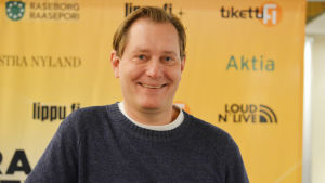 Medieprofilen Petteri Ahomaa är har jobbat som nyhetsankare på yle, men är nu producent för mtv3:s Uutisvuoto. En bild på en man, Petteri Ahomaa, som står framför en duk som gör reklam för en festival.