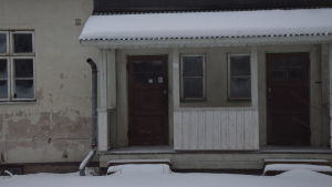 Det här vitrappade huset har också haft hyresgäster, men det är ett bra tag sedan. Den senaste hyresgästen var en förening. Ett vitrappat hus där rappningen delvis fallit bort och ett stuprör hänger också löst. Vinter och snö. Huset är obebott.