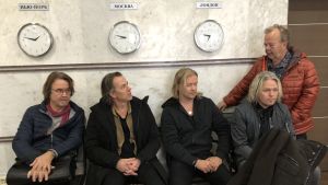 Johan Becker (tredje från vänster) tillsammans med de övriga medlemmarna i Secret Service. Fem medelåders män sitter på en bänk under tre klockor som visar olika tider. Ryska skyltar syns ovanför klockorna.