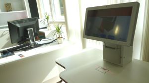 I ett meröppet bibliotek kan man låna böcker även då personalen inte är på plats. En låneautomat, en dator som man visar biblioteksbokens streckkod för för att låna den. I bakgrunden ett arbetsbord med vanlig bokdator. Solen lyser in i rummet. Bibliotek.