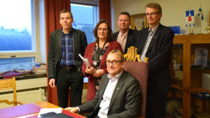 Denis Strandell (sittande i stolen) och hans kollegor i ledningsgruppen: Juha Kuittinen, Elisabeth Kajander, Karl-Erik Gustafsson och Jukka Takala. Hangös stadsdirektör Denis Strandell sitter i en stol vid ett bord. Bakom honom står Juha Kuittinen, Elisabeth Kajander, Karl-Erik Gustafsson och Jukka Takala.