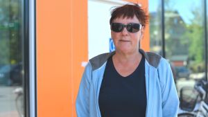 Viveca Houtsonen Närbild, porträtt, av Viveca Houtsonen, en kvinna med mörkrött kort hår, solglasögon och ljusblå huvtröja utanför K-market i Hangö.