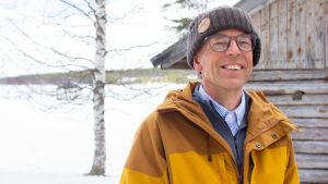 Klikkaa kuvaa ja lue lisää! Avaruusfyysikko Esa Turunen keltaisessa ulkoilutakissaan, harmaan vajan edessä. Taustalla näkyy jäinen joki.