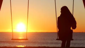Känslan av att vara ensam och isolerad kan vara traumatisk. Kvinna sitter på gunga i solnedgång, bredvid henne en tom gunga