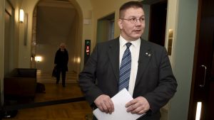 Riksåklagaren utreder om Juha Mäenpää bör åtalas för hets mot folkgrupp på grund av hans uttalanden. Men först behandlar grundlagsutskottet frågan om riksåklagaren får väcka åtal för uttalanden som kan anses som hets mot folkgrupp. En man med glasögon och kostym i riksdagens korridor.