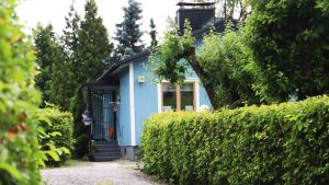 Husen ritades i Finland men byggdes färdiga av element i Sverige. En grusväg leder fram till ett blått hus skymtar bakom grönskande träd och buskar.