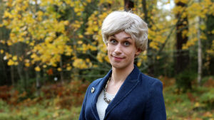 Christoffer Strandberg eller Elisabeth Rehn? Christoffer Strandberg utklädd till Elisabeth Rehn. Han bär en kort blond peruk, blå kavaj med en brosch och ett halsband.