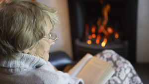 De flesta äldre vill bo kvar hemma så länge som möjligt, visar flera utredningar. En äldre kvinna sitter och läser en bok framför en öppen spis.