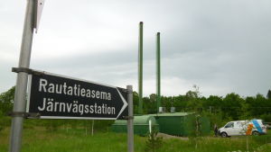 Advens fjärrvärmeverk. Tåg i stället för bil och biobränslen i stället för fossila. Så kan Sjundeåbor minska på klimatusläppen. En skylt med texten "Rautatieasema Järnvägsstation". I bakgrunden ett litet grönt fjärrvärmeverk med två skorstenar, en paketbil står utanför värmeverket. Grönt gräs.