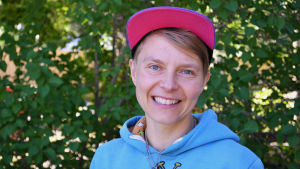 Virve Savoila. En bild på en kvinna med rosa keps och blå tröja som står framför en buske. Personen på bilden är Virve Savoila