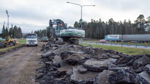 Just nu pågår arbetet med att ta bort den gamla asfalten. En grävmaskin lastar uppbruten asfalt på en lastbil.