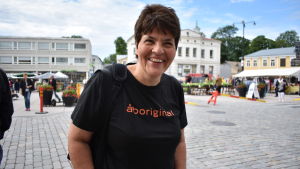Lotte Rasinkangas är på besök från Åbo. Kvinna med svart skjorta och mörkt hår ser mot kameran och ler. Torg i bakgrunden.
