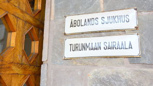 Bergendahl intygar att alla vid Åbolands sjukhus kan tala svenska, men han anser ändå att fördjupade kurser i språket hela tiden är nödvändiga. en vit skylt framför en stenvägg bredvid en trädörr