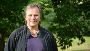Jan Jacke Jacobson. porträttbild av jan jacobson. han står ute i grönskan och bär en svart jacka över en lila t-skjorta.