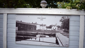 Dockan vakar över det gamla fotografiet av Fisktorget. Ett staket med ett gammalt, uppförstorat fotografi och en docka som kikar över staketet.