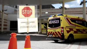 Kuvassa etualalla näkyy kyltti jossa kehoitetaan koronanäytteenottoon tulevia odottamaan ja taustalla näkyy ambulanssi.