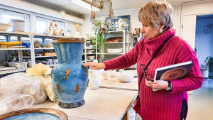 Just nu inspireras Åsa Hellman av Medelhavet. Det vittnar havsmotiven och den speciella blå färgen om. Glasyr med vacker guldlyster finns i många av Hellmans verk. En kvinna i pink tröja står intill ett bord med keramikgföremål på. Hon sträcker ut handen och rör vid en blå kruka. I famnen håller hon en bok och sina glasögon.