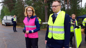 Åklagaren Marianna Semi och försvarsadvokaten Mikko Puhakka på olycksplatsen. En kvinna med ljusröd väst och en man med gul väst utomhus på en skogsväg.