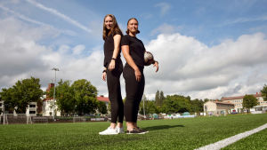 Felicia Gröning och Jill Illman är två av drygt 130 tjejer och kvinnor som spelar i den grönvita EIF-skjortan. Felicia Gröning och Jill Illman står rygg mot rygg på en konstgräsplan. Jill håller i en fotboll.