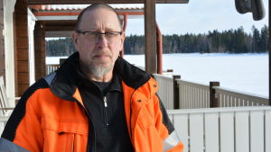Kenneth Härtull. en porträttbild av kenneth höglund. han står utomhus med sin veranda i bakgrunden. han bär glasögon och en orange arbetsjacka.