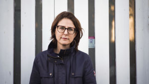 När Ira Frey-Kajanders dotter inte känner sig trygg med daghemspersonalen på grund av att de är för få så står hon och väntar på sin mamma vid daghemmets port. En besämd kvinna tittar åt vänster. I bakgrunden en dörr med ljusblå träplankor och avlånga glasfönster.