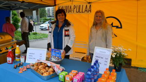 Jill Öhman och Maarit Rope. Två kvinnor står inne i ett gult torgtält bakom ett bord med olika drycker och sötsaker till salu.