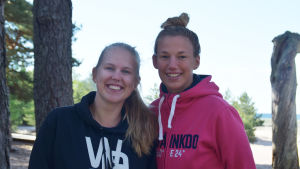 Glada lägerledare som nyligen vaknat upp efter första natten på Breidablick för den här sommaren. Både Ida Hultgren och Emmi Wessman tillbringar sin andra sommar som lägerledare i Hangö. En bild på två personer som ler mot kameran. I bakgrunden syns tallar, sand och hav.