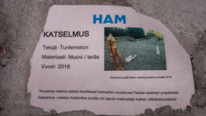 Alldeles intill statyn finns den här informationskylten. Informationsskylt om ett konstverk. Skylten är fäst i betong.