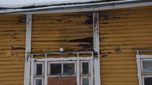 Flyg fågel flyg, men helst inte in. Hålen i fasaden har täckts med metallnät. En gul trävilla, gammal arbetarbostad med fler bostäder. Målfärgen flagar, stockarna är ruttna vilket lett till hål i ytterväggarna.