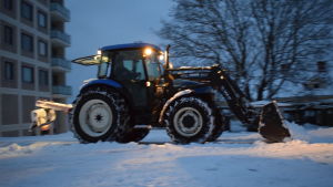 Snöplogarnas sikte är ofta dåligt då de plogar. En traktor som plogar snö en tidig vintermorgon. Strålkastarna på traktorn lyser.