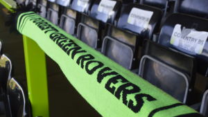 Förenta Nationerna har uppmärksammat Forest Green Rovers som det enda koldioxidneutrala fotbollslaget i världen. En grön halsduk med texten Forest Green Rovers i svart spänd över ett räcke framför ihopvikta bänkar på en läktare.