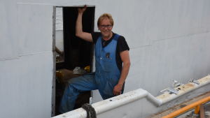 Samuli Penttinen är ingenjör och jobbar flitigt med att renovera båten. En man i blå overall står i en dörröppning på en vit metallbåt.