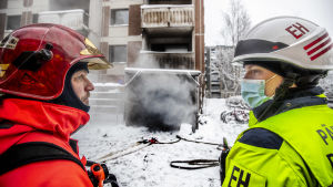Eemu Hyvönen (till höger) diskuterar med en rökdykare hur räddningsarbetet ska utföras. Två brandmän, en i röda skyddskläder och den andra i gula, och med skyddshjälmar pratar med varandra utanför ett höghus därifrån det bolmar ut svart rök.