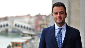 Simone Venturini är ansvarig för turismen i Venedig. Han är betydligt yngre än de flesta italienska politiker både på lokal och nationell nivå i landet. En man med kort mörkt hår, mörk kostym, ljusblå skjorta och vitprickig blå kravatt ler mot kameran. I bakgrunden suddigt en bro i Venedig.