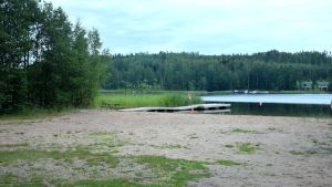 Mattvätt får inte ligga bredvid badstranden. Badstrand vid havsvik med brygga och träd intill. Vass ute i vattnet och på andra sidan syns båtar, hus och träd.