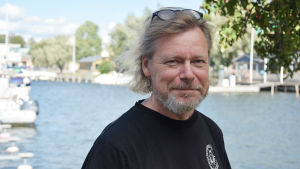 Marko Reinikainen är direktör för Tvärminne zoologiska station vid Helsingfors universitet. En man står i svart t-skjorta och ler mot kameran. I bakgrunden syns en hamn med vatten och båtar. Även träd syns i bakgrunden.