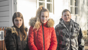 Kaja Čolić (t.v), Lukas Savković och Jovala Źurovac säger att det är roligt att vara på besök i Finland. Tre barn står framför ett vitt trähus. De ler och ser in i kameran.