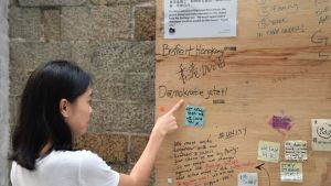 Naomi Wong pekar på en så kallad Lennon vägg nära sitt hem. Anslagstavlorna har fått sitt namn efter John Lennon, och på dem kan man fästa budskap om demonstrationerna. Kvinna pekar på plakat i trä. Ordet demokrati står skrivet på plakatet.
