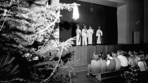 Julfest i Aleksis Kiven peruskoulu år 1969. En svartvit bild på lågstadiebarn utklädda till stjärngossar som står på en scen på skolans julfest. Barn sitter i publiken och tittar på.
