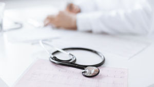 En läkares händer ligger på ett bord. I förgrunden ligger ett stetoskop och ekg-utskrifter.