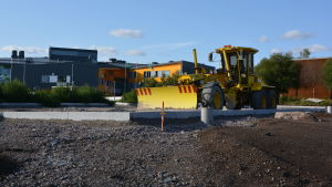 Det här ska bli Stora Byvägens anslutningsparkering. I bakgrunden syns en del av Nickby Hjärta. Där håller man också på och bygger eftersom skolcentret ska utvidgas. En gul traktor står på en sandplan. I bakgrunden syns Nickby Hjärta.