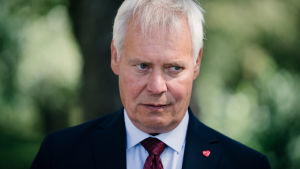Statsminister Antti Rinne tror att arbetsmarknadsparterna har intresse av att påverka hur regeringen skapar nya arbetsplatser. Antti Rinne i mörk kostym och röd slips mot en grön suddig bakgrund. Han har en sammanbiten min.