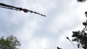 ... högt upp i luften, utan att skada träden runt huset. En lyftkran fotad nerifrån. Ser mest grå himmel.