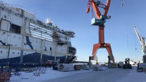 Aurora Botnia vid varvet i Raumo. Halvfärdigt fartyg vid kaj intill en orange lyftkran. Det finns snö på marken och himlen är blå.