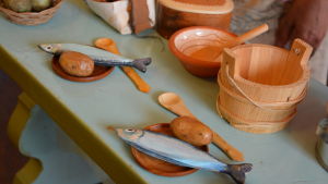 Fiskar gjorda av trä och potatisar gjorda av tidningspapper kan man hitta på miniatyrborden för barn. På en bänk ligger två tallrikar med konstgjord fisk och potatis, i bakgrunden står det kärl gjorda av trä.